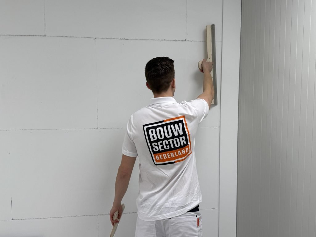 Stukadoor in wit werkpak strijkt met een lange spaan over een gipswand, van achteren gezien