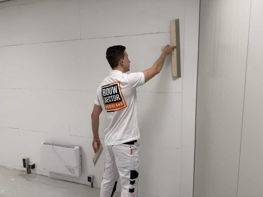 Stukadoor reikt met een lange spaan hoog tegen de wand, vlak onder het plafond
