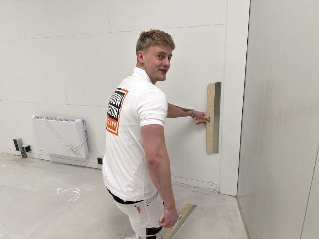 Lachende stukadoor zet een spaan tegen een wand in een lege ruimte met een radiator